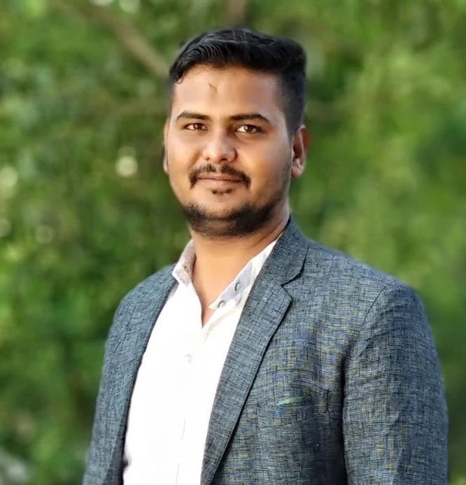 Portrait of Tanveer Alam, wearing a grey checked jacket over a white shirt, photographed outdoors against foliage.